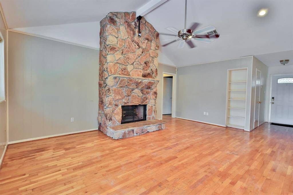 This spacious living room features a striking stone fireplace, hardwood floors, and a vaulted ceiling with a ceiling fan, creating a warm and inviting atmosphere. Built-in shelving adds functionality, and the room is filled with natural light.