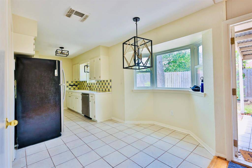 This photo features a bright kitchen with white cabinetry and a modern geometric light fixture. The space includes a tiled floor, black refrigerator, and a scenic bay window overlooking the backyard. The backsplash adds a colorful touch, complementing the overall airy ambiance.