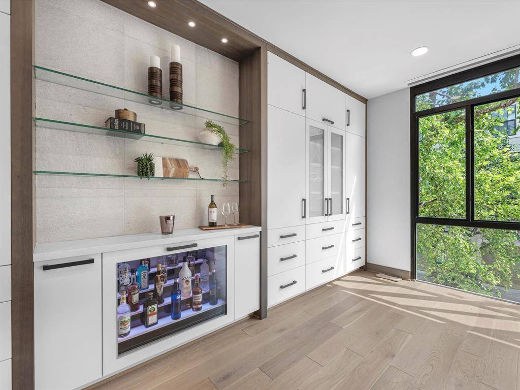 This wall features a classy, lit drinks fridge and an additional wine fridge. The space is bright and airy, thanks to a large window that offers a view of lush greenery outside.