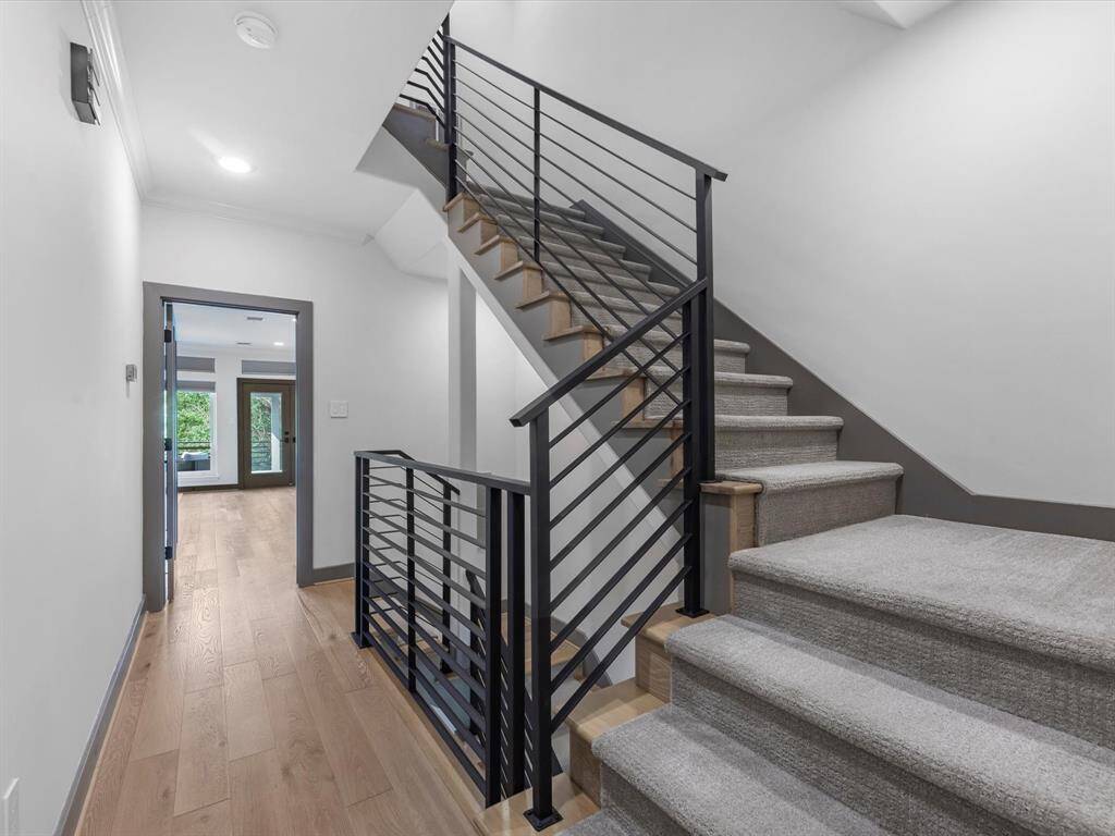 The sleek modern staircase features metal railings and carpeted steps. The hardwood floor leads to a room with large windows and a glass door, offering a view of greenery.
