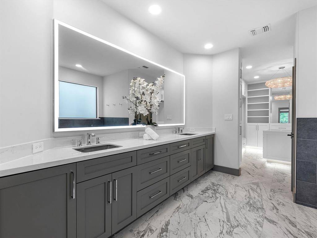 It features a modern design with a large, backlit mirror above a double vanity with sleek gray cabinets. The marble floor adds elegance, and there's ample natural light from a frosted window.