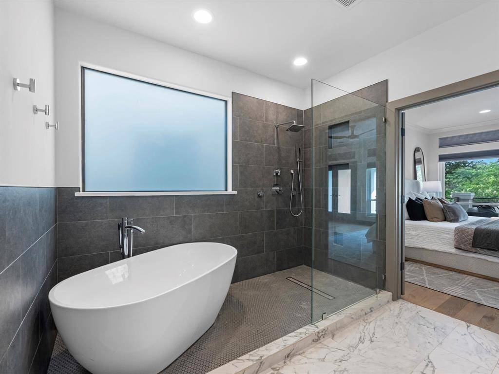 The modern bathroom features a sleek freestanding bathtub, a spacious glass-enclosed shower with dark tile, and a large frosted window for natural light. It connects directly to the primary bedroom, enhancing convenience and flow.