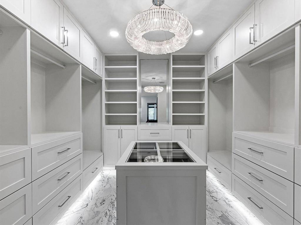 Just WOW! The walk-in closet is spacious and elegant with ample shelving and drawer space. It features a central island with glass top and is beautifully lit by a modern chandelier and recessed lighting, all set against a sleek, white finish and marble flooring.