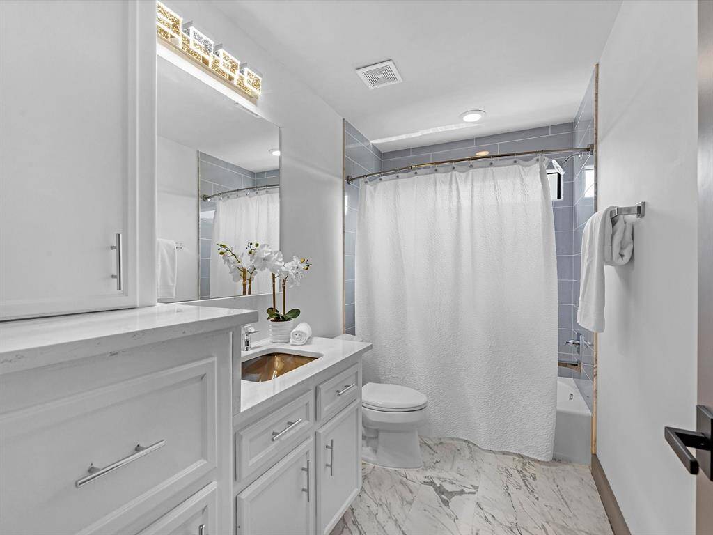 The bathroom as per the rest of the home, features a clean, modern design with white cabinetry and marble-patterned flooring. It includes a large mirror, ample lighting, and a shower-tub combination. The space is bright and elegantly finished with decorative touches.