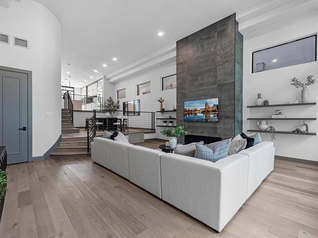This modern living space features an open floor plan with sleek wooden flooring, spectacular fireplace. The room is bright and airy, enhanced by large windows, contemporary shelving, and a staircase leading to the dining room.