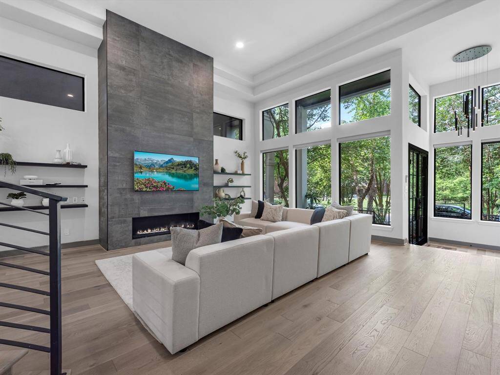 This modern living room features high ceilings and expansive windows providing abundant natural light and serene views. A sleek fireplace and included TV are set into a striking, minimalist wall, on a warm wood floor. Perfect for stylish, open-concept living.