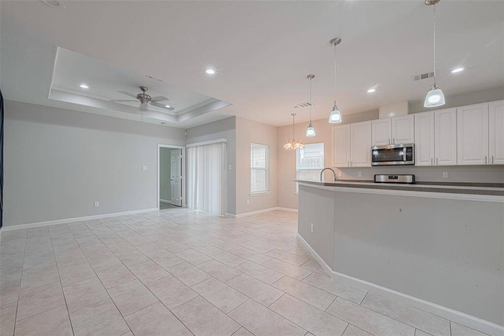 Showcase a spacious open floor plan living area with a modern kitchen featuring white cabinetry, stainless steel appliances, and pendant lighting. The room has a tiled floor, recessed ceiling with a fan, and ample natural light through large windows and a sliding glass door. Ideal for entertaining or family living.