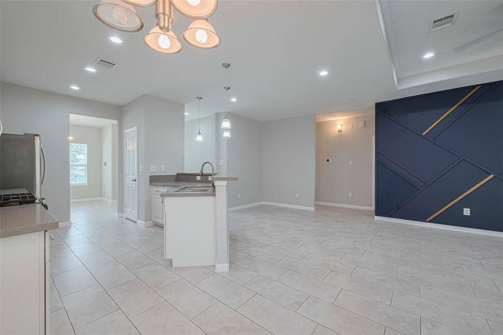 This open-concept living area features a modern kitchen with stainless steel appliances and a central island. The space is bright with recessed lighting and a unique accent wall, complemented by light tile flooring.
