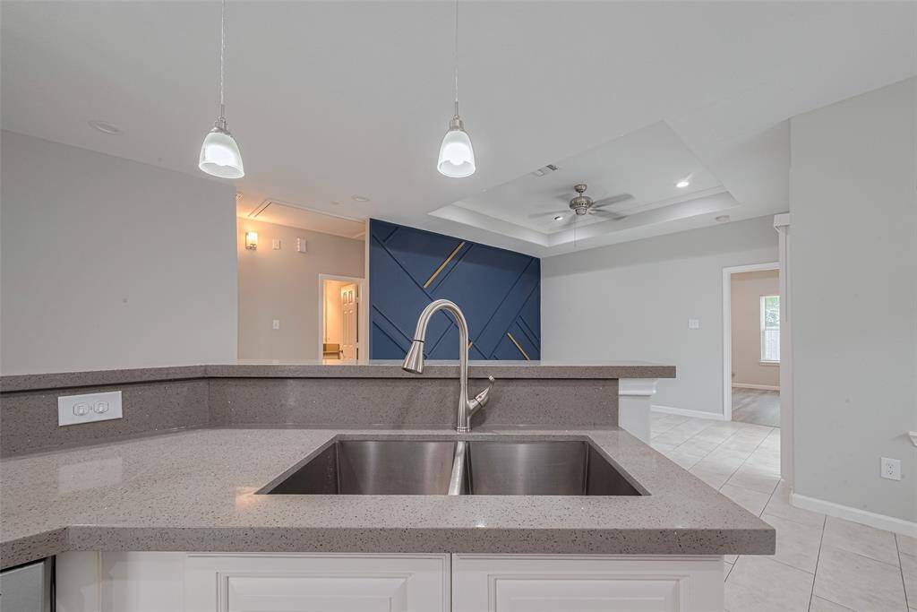 Modern kitchen features include, dual-basin sink with a stylish faucet on a spacious quartz countertop. Pendant lighting adds elegance, while the open layout connects to a living area with a unique accent wall and ceiling fan, creating an inviting atmosphere.
