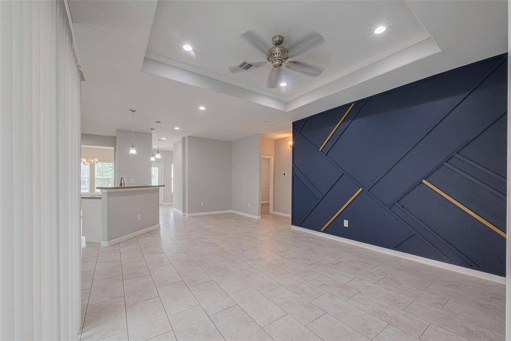 The spacious, open-concept living area features a modern design with a striking accent wall, recessed lighting, and a ceiling fan. The neutral tile flooring extends to the kitchen, which has a breakfast bar and pendant lighting. Perfect for contemporary living.
