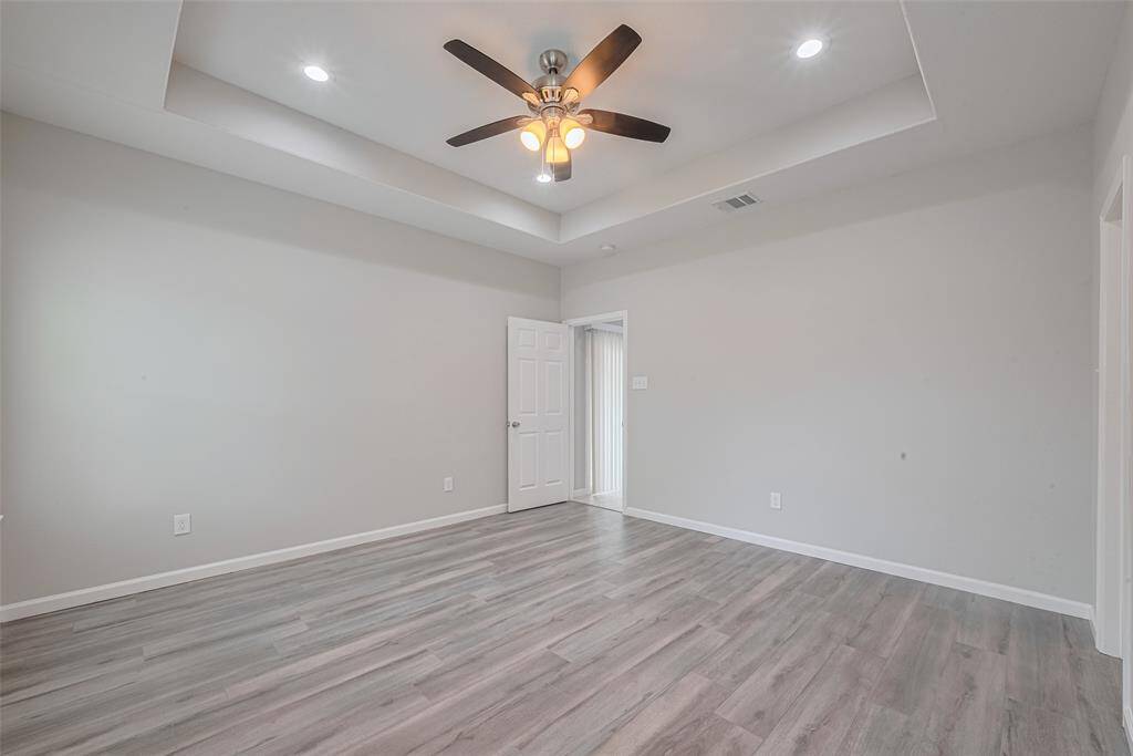 Spacious main bedroom features modern gray flooring, a tray ceiling with recessed lighting, and a ceiling fan. The neutral walls provide a clean, fresh look, while a door leads to an adjacent space, offering versatile use.