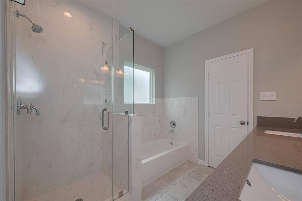 The bathroom features a modern design with a glass-enclosed shower, a separate bathtub, and a double vanity, his and hers. The neutral color palette and large window provide a bright, airy feel.