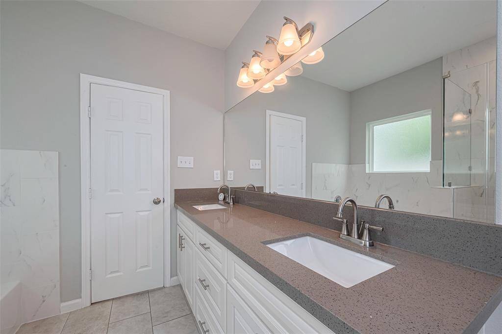 The bathroom features a modern dual-sink vanity with a large mirror and stylish lighting. The room has a neutral color palette, a frosted window for natural light, and a separate shower area with tiled walls.