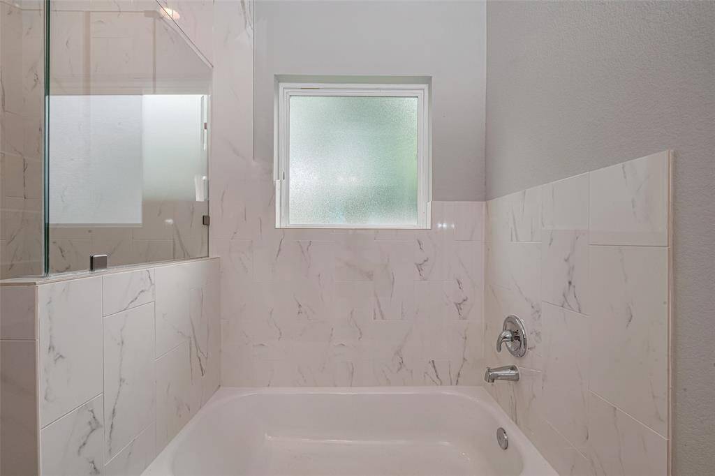 The bathroom features a modern tub with elegant marble-look tiles, a frosted window for privacy, and a sleek chrome faucet.