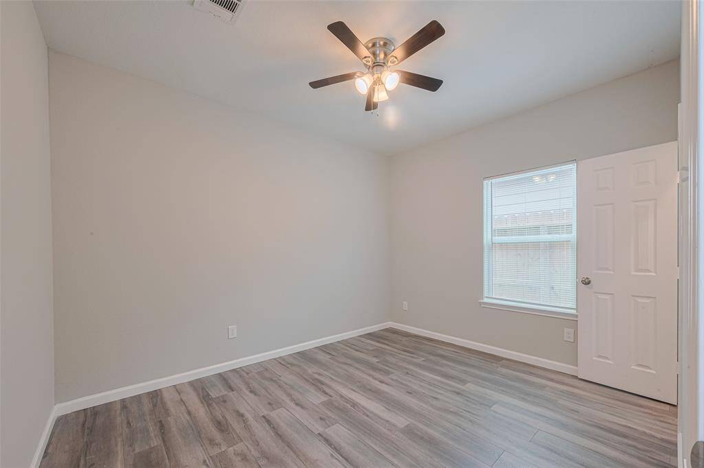 The second bedroom features a modern, neutral color scheme with light gray walls and wood-style flooring. It has a ceiling fan with lights, a window providing natural light, and a standard white door. Perfect for a cozy bedroom or home office.