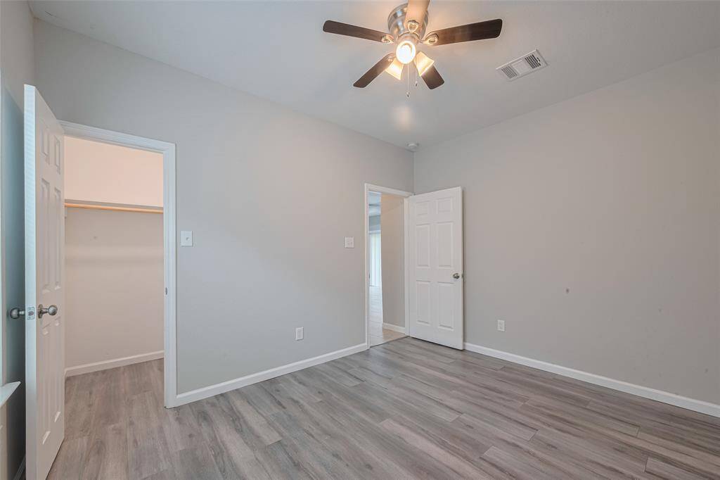 Another look of the secondary bedroom features a neutral color palette with light gray walls and laminate wood flooring. It includes a ceiling fan, a closet with shelving, and a white door leading to other areas of the home.