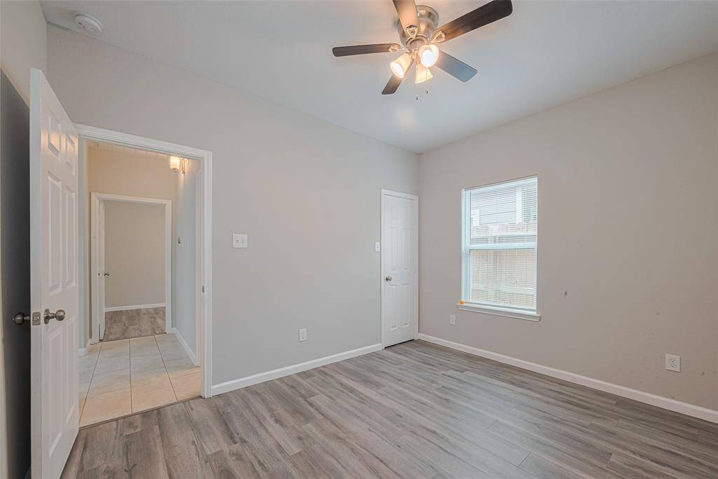 The third bedroom features light gray walls, wood-style flooring, and a ceiling fan with lights. It has a window for natural light and an adjoining hallway with tiled flooring.