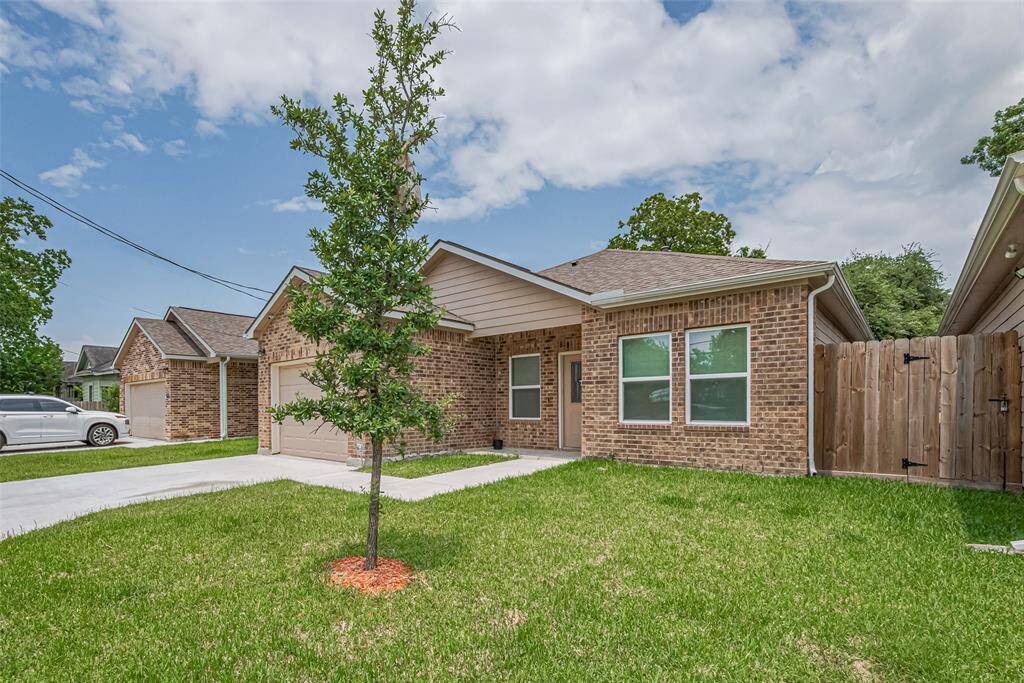 This photo features a charming, single-story home with a neatly maintained front lawn. The property includes a driveway leading to an attached-detached garage and a wooden fence providing privacy. The exterior is complemented by a tree and clear blue skies.
