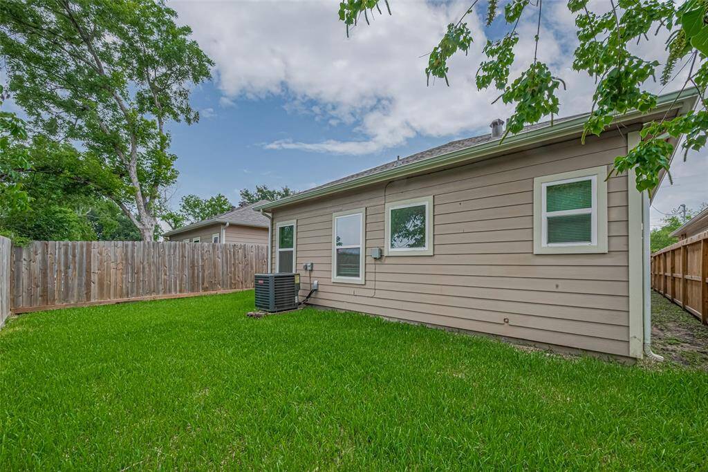 The single-story home's exterior with beige siding, two windows, and a neatly maintained backyard. The yard is enclosed by a wooden fence, providing privacy, and there's an HVAC unit visible against the house. Lush green grass and surrounding trees add a touch of nature.