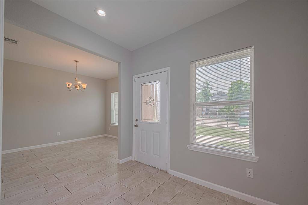 This photo features a bright entrance area with a tiled floor, neutral walls, and a window allowing natural light. It opens into a dining space with a chandelier, creating an inviting atmosphere.