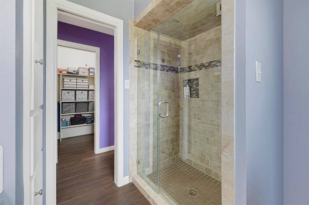 A glimpse of the beautifully tiled walk-in shower, featuring elegant glass tile inlays, frameless glass doors, recessed shelving, and a walk-in closet just beyond.
