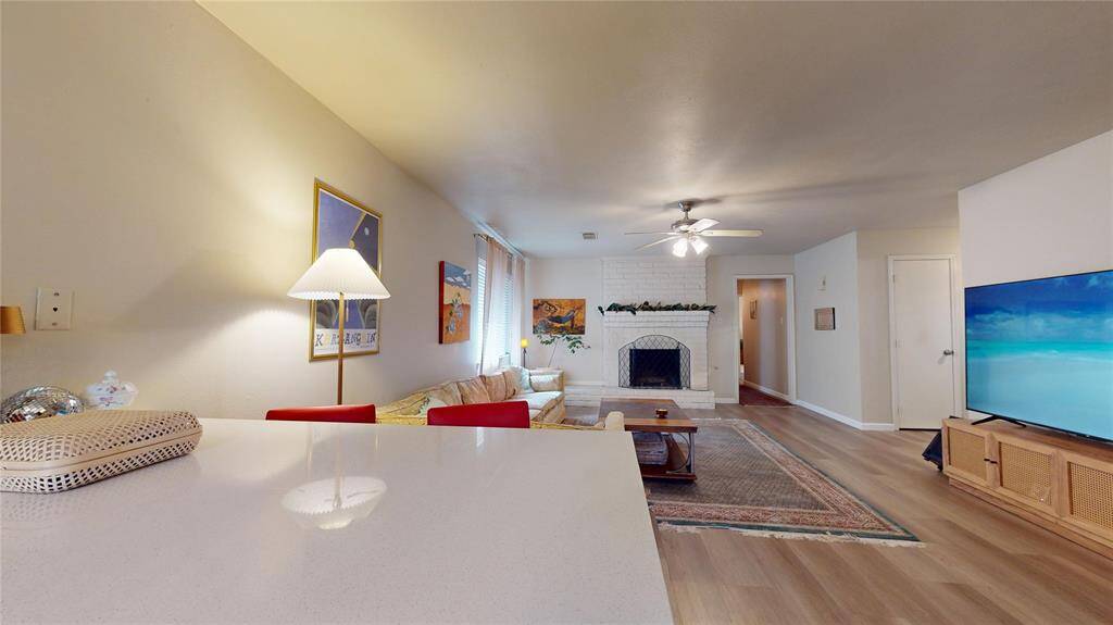 This cozy living room features a neutral color palette with a white brick fireplace, modern ceiling fan, and ample natural light. It includes a large TV, comfortable seating, and stylish decor, creating a welcoming atmosphere.