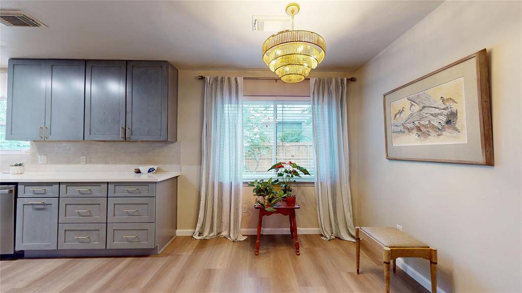 This photo showcases a bright and stylish kitchen area with modern gray cabinets, a chic chandelier, and a large window with sheer curtains. The breakfast area features light wood flooring and is accented with framed artwork and a small plant, creating a cozy and inviting atmosphere.