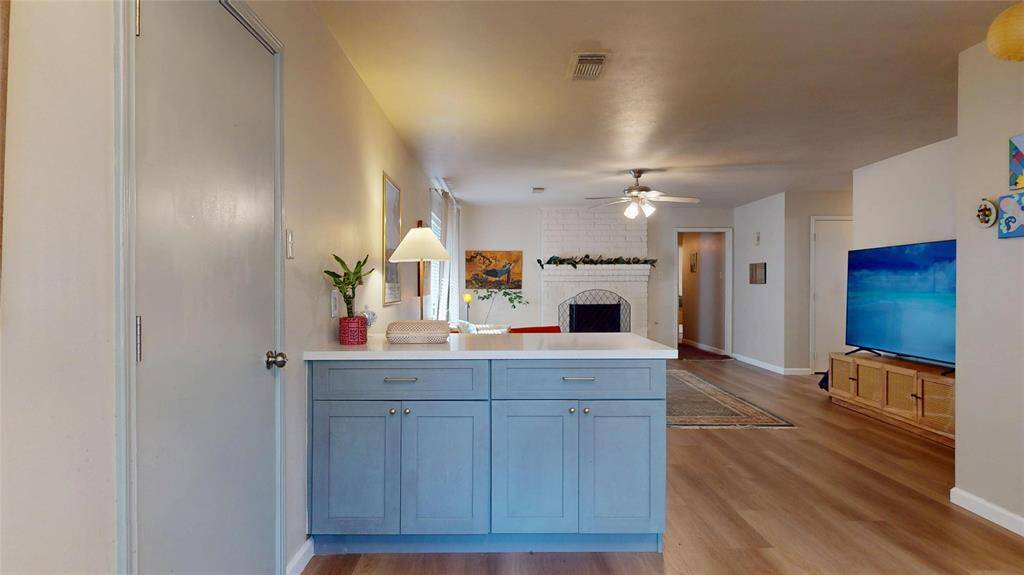 This photo showcases a cozy living space with hardwood floors, a white brick fireplace, and a ceiling fan. There's a blue cabinet with a lamp and a plant, and a wall-mounted TV on a wooden stand. The room feels open and inviting.