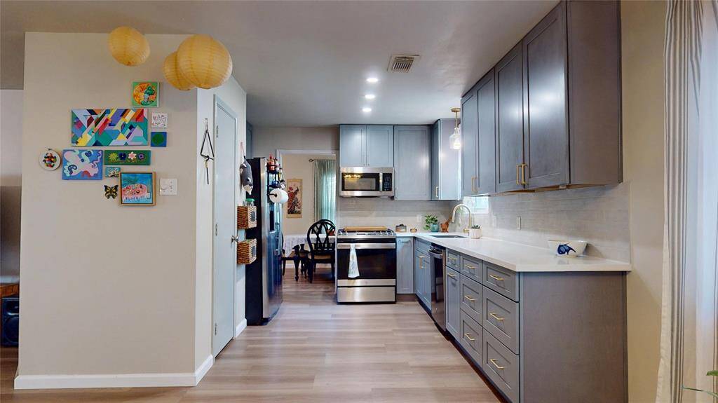 This kitchen features sleek gray cabinets, modern stainless steel appliances, and a bright, open layout. The light wood flooring and ample counter space create a welcoming atmosphere. Decorative accents add a personal touch, making it both stylish and functional.