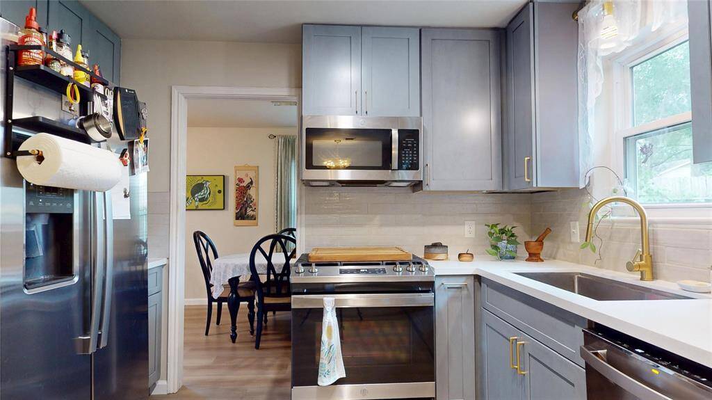 This kitchen features modern gray cabinets, stainless steel appliances, and a sleek countertop with a gold-tone faucet. It has a window for natural light and an open doorway leading to a dining area, creating a cozy and functional space.