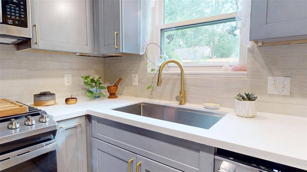 This kitchen features modern gray cabinetry with brass hardware, a sleek stainless steel sink with a stylish gold faucet, and a gas stove. The light countertop and backsplash, combined with natural light from the window, create a bright and inviting space.