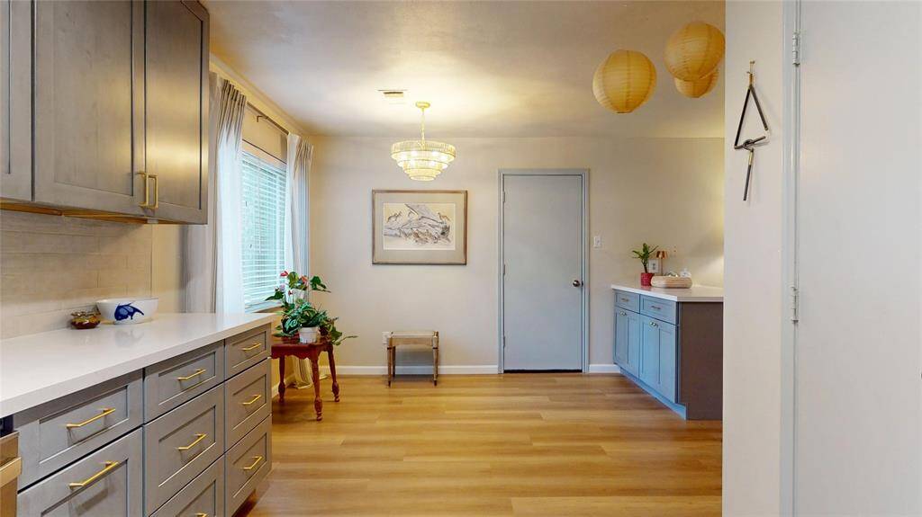 This photo showcases a bright kitchen area with modern grey cabinetry, gold hardware, and sleek white countertops. The space features a warm wood floor, a large window with curtains, decorative lighting, and a small side table with plants, creating a cozy and stylish atmosphere.