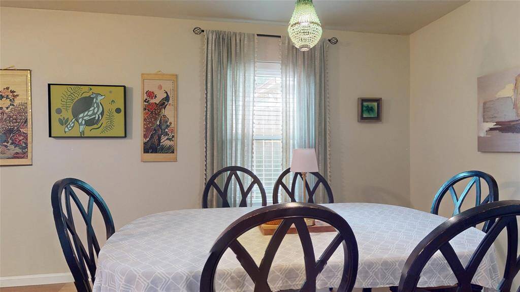 This dining room features a round table with elegant black chairs and a light tablecloth. The room is brightened by a window with soft curtains and a hanging light fixture. The walls are adorned with various artworks, adding character and warmth to the space.