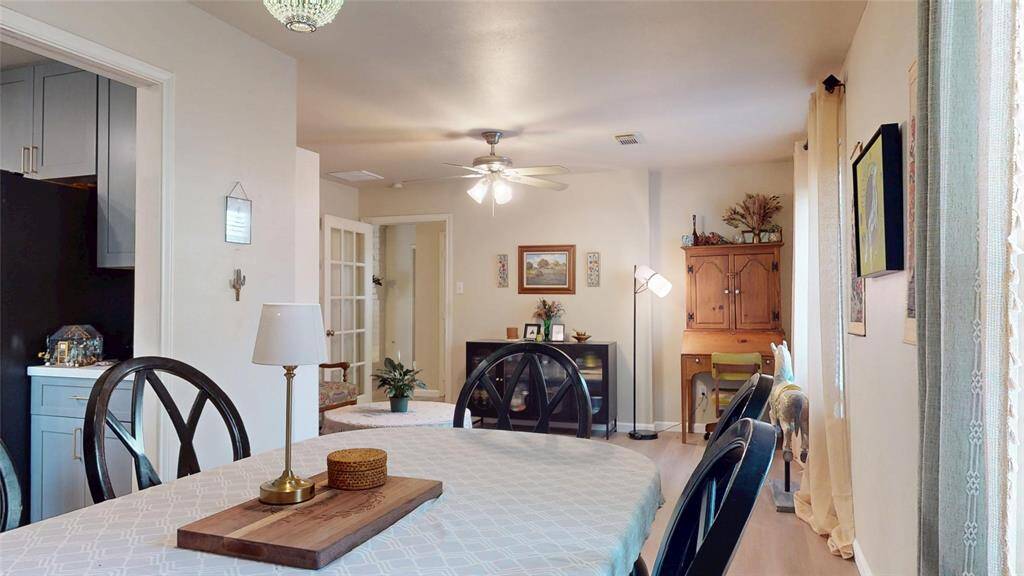 This photo showcases a cozy dining area with a table and chairs, leading into a bright living space. The room features warm, neutral tones, a ceiling fan, and natural light from a window. Decorative elements include wall art and a small wooden desk. The space feels inviting and functional.
