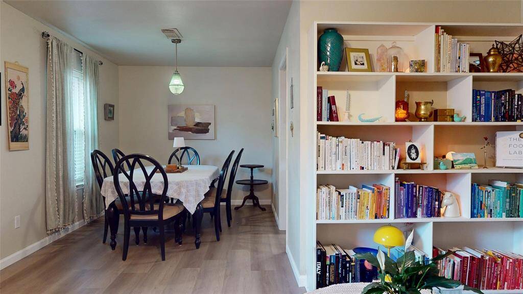 This cozy dining area features a table with elegant dark chairs, a soft pendant light, and light-colored walls. Adjacent is a built-in bookshelf filled with books and decorative items, adding character and warmth. Large windows with curtains offer natural light, creating an inviting atmosphere.