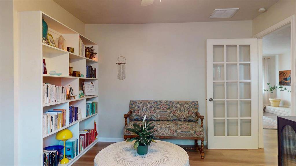 Cozy reading nook featuring a built-in bookshelf filled with books and decor. The room includes a patterned two-seater chair, a round table with a plant, and a French door leading to a bright adjoining space.