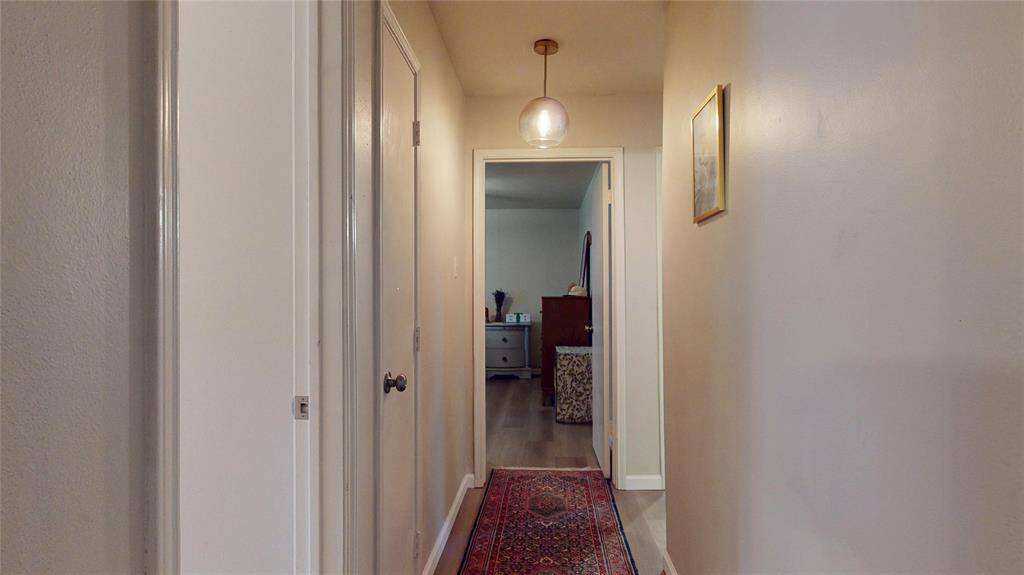 This photo shows a cozy hallway with light-colored walls, a hanging pendant light, and a patterned rug leading to a room. There's a framed picture on the wall and a glimpse of furniture in the adjoining room, suggesting a warm, inviting space.