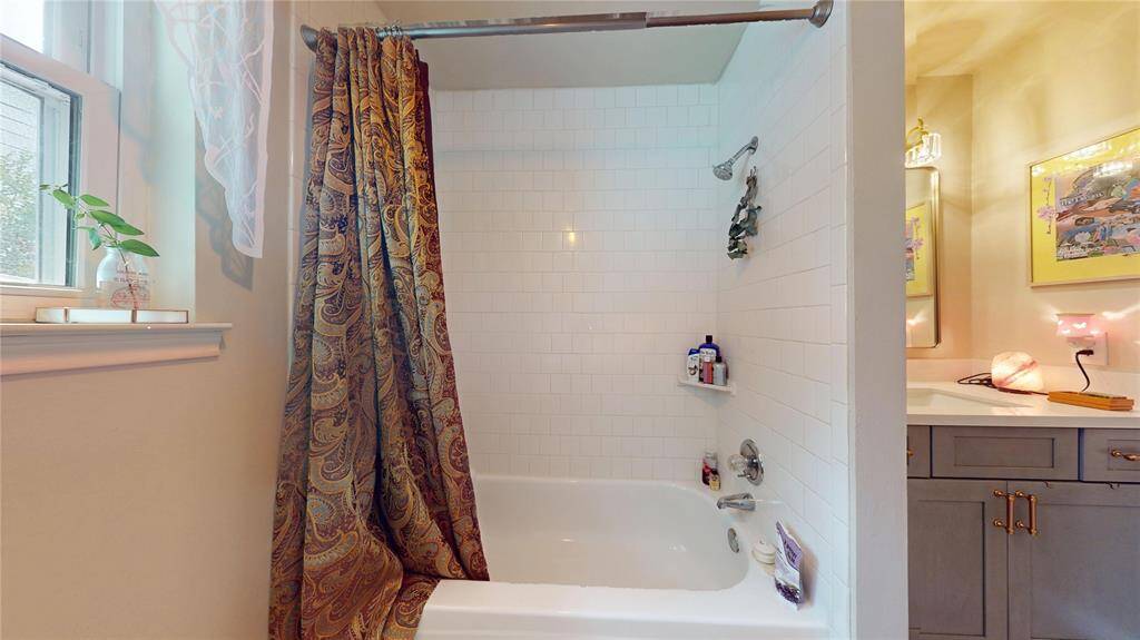 This cozy bathroom features a classic white tiled shower and tub, a decorative curtain, and a bright, inviting atmosphere. A nearby window allows natural light to fill the space, complemented by a stylish vanity with ample storage.