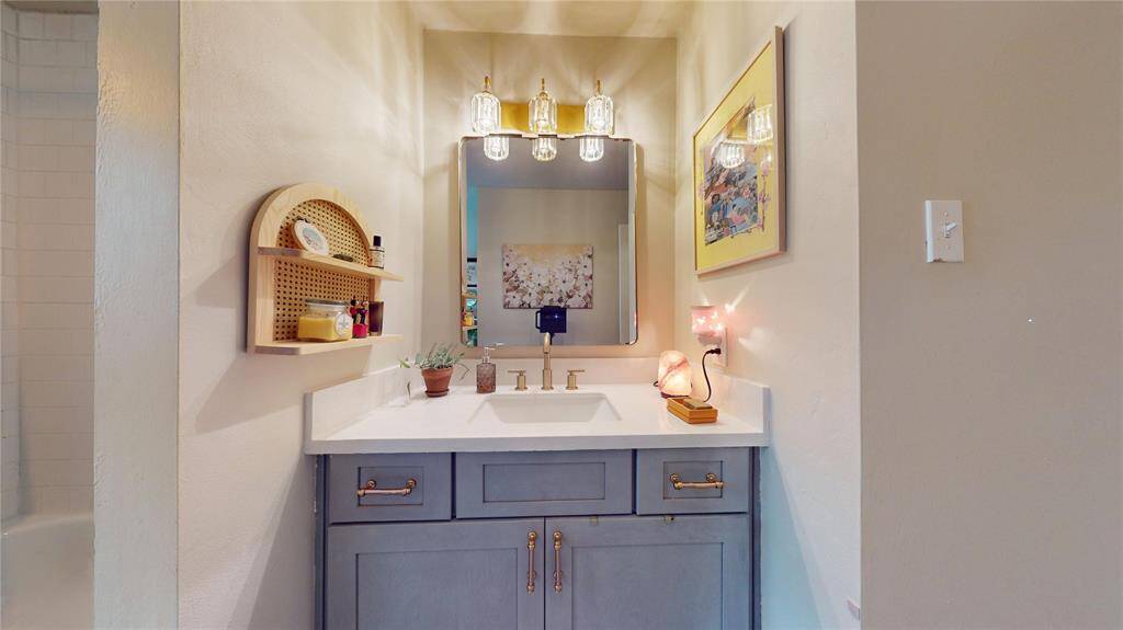 This bathroom features a modern vanity with a white countertop and stylish brass fixtures. It has a framed mirror with elegant lighting above, and a small shelf for toiletries. The neutral walls are complemented by decorative artwork, creating a cozy and functional space.