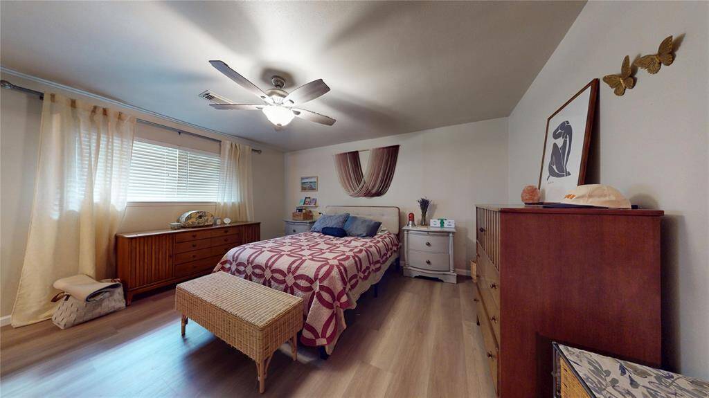 This cozy primary bedroom features a double bed with a stylish quilt, a ceiling fan, and natural light from a large window with curtains. It includes wooden furniture such as dressers and a wicker bench, and has decorative art on the walls, creating a warm and inviting atmosphere.