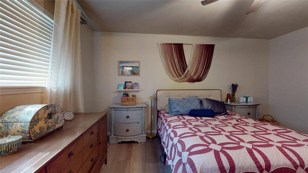This cozy primary bedroom features a comfortable bed with a decorative quilt, a wooden dresser, and a bedside table. Natural light streams in through a window with blinds and curtains, creating a warm ambiance. The room is tastefully decorated with a wall hanging and small art pieces.