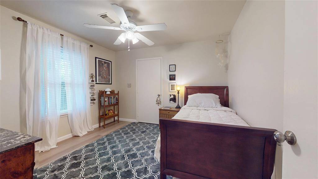 Cozy bedroom featuring a single bed, ceiling fan, and natural light from a large window with sheer curtains. The room includes a small bookshelf, decorative artwork, and a patterned area rug, offering a comfortable and inviting space.