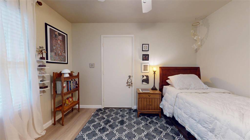 This cozy bedroom features a single bed with a wooden headboard, a bedside table with a lamp, and a decorative rug. There's a small wooden shelf for storage and wall art for a personal touch. Natural light filters through sheer curtains, creating a warm, inviting atmosphere.