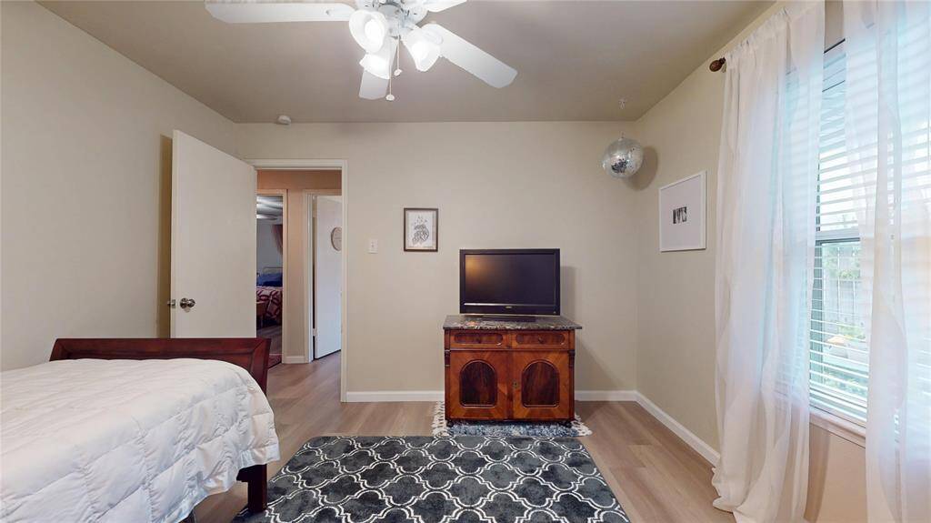 This room features neutral walls, a ceiling fan, and a window with sheer curtains allowing natural light. It includes a single bed, a TV on a wooden stand, and a patterned rug, creating a cozy, functional space.