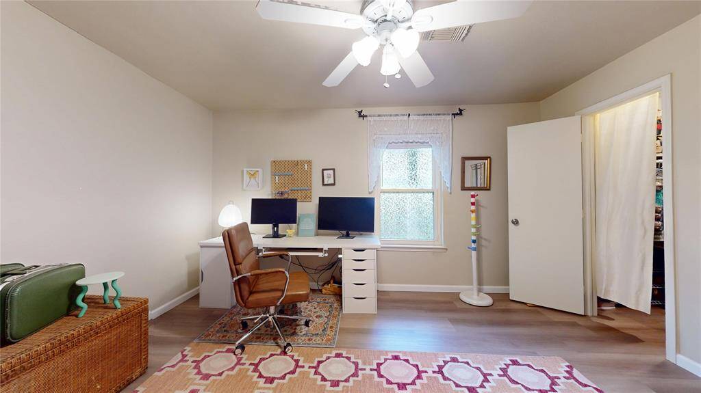 This is a cozy bedroom with neutral walls, featuring a desk with dual monitors, a comfortable chair, and a ceiling fan for comfort. The room has plenty of natural light from a window, and a closet for additional storage.