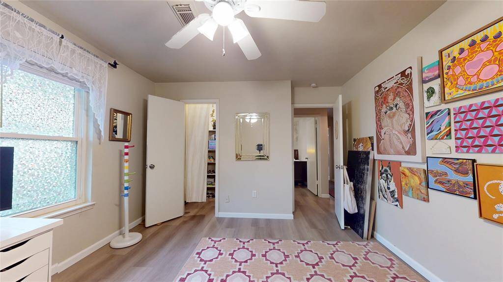 This bedroom features bright natural light from a large window, modern wood flooring, and a ceiling fan. It has a cozy atmosphere with a decorative rug and an art-filled accent wall, ideal for a creative space or home office.