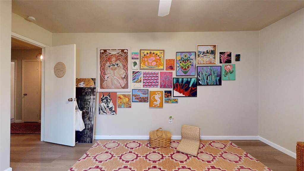 This bedroom features a neutral wall adorned with an eclectic gallery of colorful artwork. The light wood flooring complements the vibrant area rug, adding warmth and character. The space is bright and open, with a ceiling fan for comfort. It's perfect for an art lover looking for a creative and inviting atmosphere.
