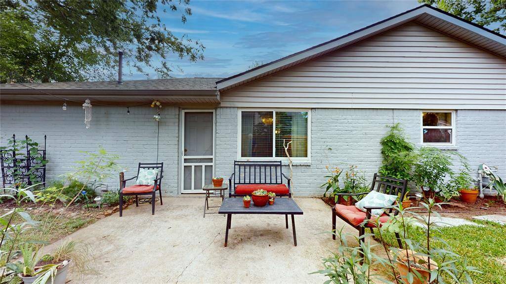 This charming patio features a cozy seating area with red-cushioned chairs and a coffee table, surrounded by lush greenery and potted plants, creating a serene outdoor retreat. The exterior is painted brick with a large window, offering a welcoming view of the home.