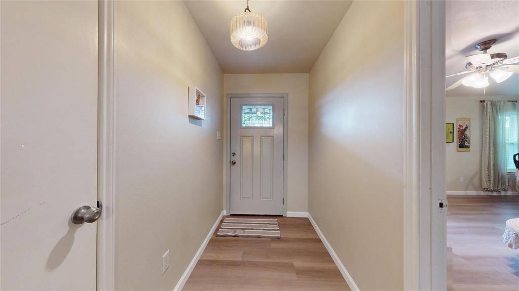This photo shows a welcoming hallway with light-colored walls and wood flooring. It features a modern light fixture and leads to a door with decorative glass. The hallway opens into a cozy living area visible on the right.