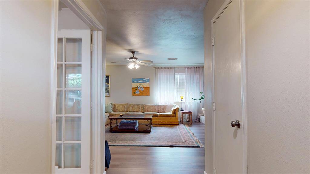 This photo showcases a welcoming living room with a cozy vibe, featuring a comfortable sofa, large windows with sheer curtains allowing natural light, a ceiling fan, and tasteful decor. The space leads from a hallway with light-colored walls and a French door, creating an open and airy atmosphere.
